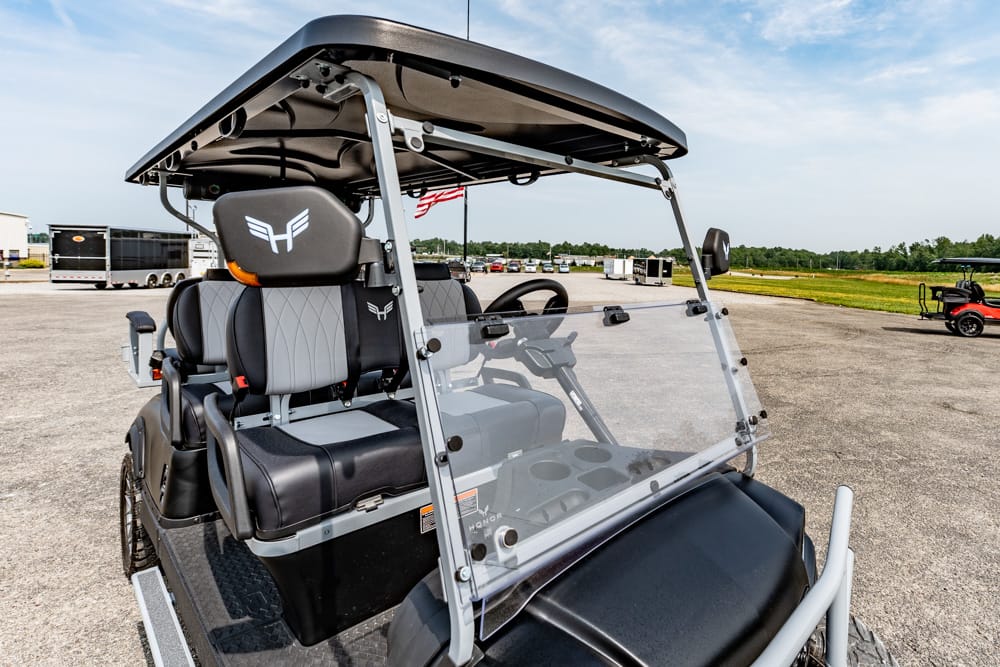 Electric golf cart with clear windshield, black and gray seats, and a canopy on a paved lot with distant trucks and an American flag in the background.