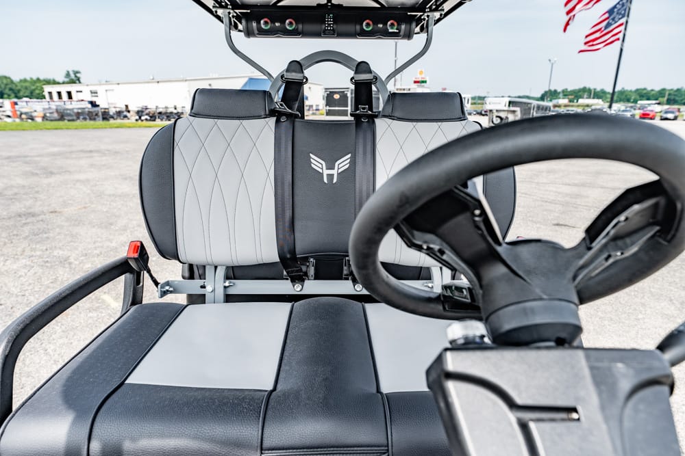 Close-up of a go-kart cockpit with a steering wheel, quilted gray seats, and a safety harness; outdoor lot in the background.