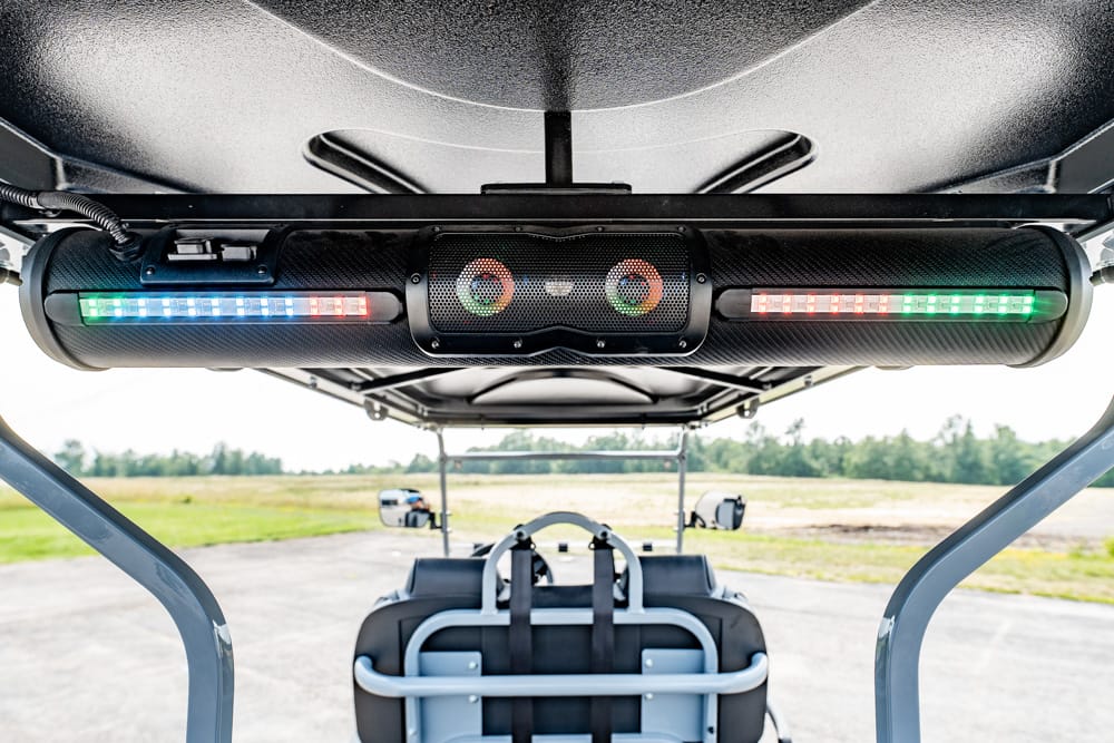 Under the chassis of a golf cart, a cylindrical LED lighting bar with multicolor LEDs (green, red, blue) is mounted, with the back seating area visible beyond.