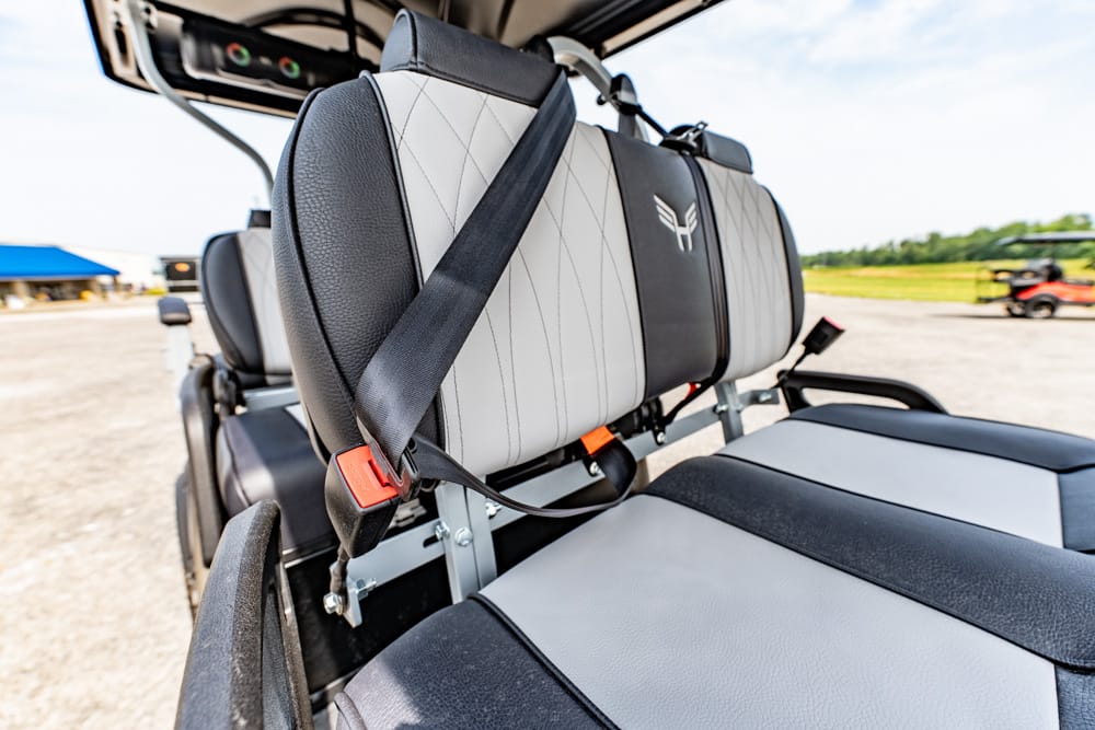 Close-up of a gray and black aircraft seat with a red seatbelt buckle in the foreground and a two-tone cushion behind it.