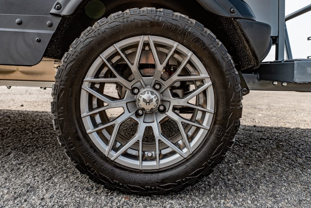 Close-up of a silver multi-spoke alloy wheel with a rugged tread tire on a vehicle surface visible behind the wheel.