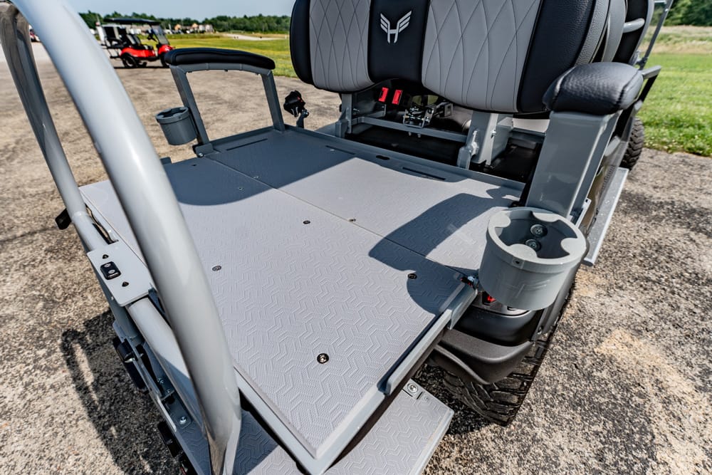 Rear cargo/platform area of a gray golf cart with a textured metal deck, cup holder, and supportive seats visible at the back of the vehicle on a sunny day