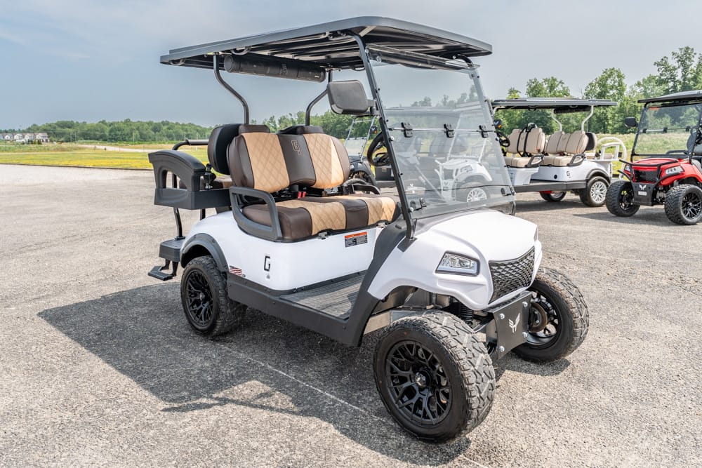 White golf cart with tan leather seats, oversized off-road tires, and a black canopy parked on a lot with other carts in the background.