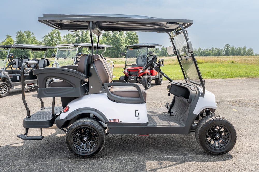 White and gray golf cart with brown seats and an open roof, parked on a paved lot with other carts in the background.