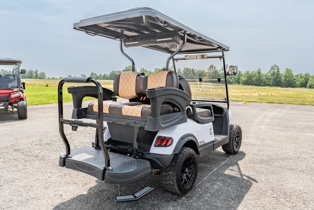 White and gray golf cart with tan seats and a rear lift, parked on a tarmac lot with fields in the background.