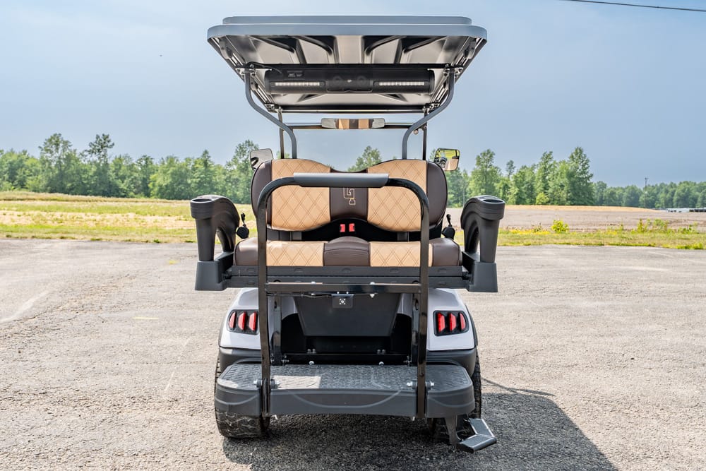 Rear view of a two-row golf cart with tan quilted seats and a black rear bar, parked on a paved lot under a blue sky.