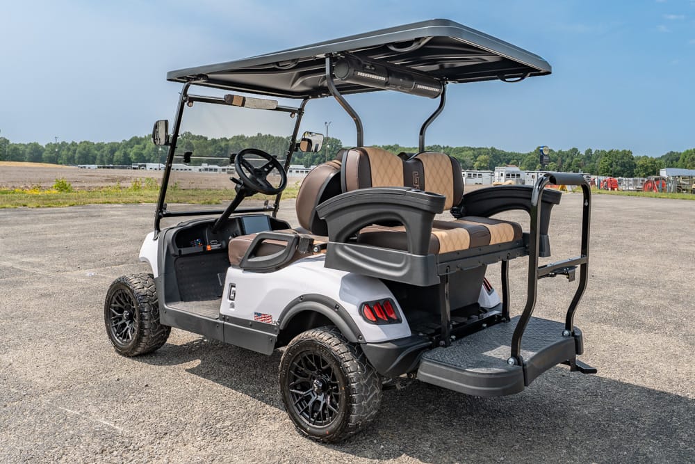 White off-road golf cart with tan seats, open sides, and a rear-facing bench on a dirt lot under a blue sky. It has a roof and rugged tires.