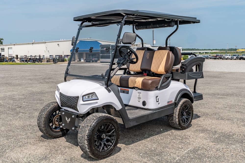 White golf cart with tan seats and black wheels parked on a gravel lot, front-left view under a canopy.