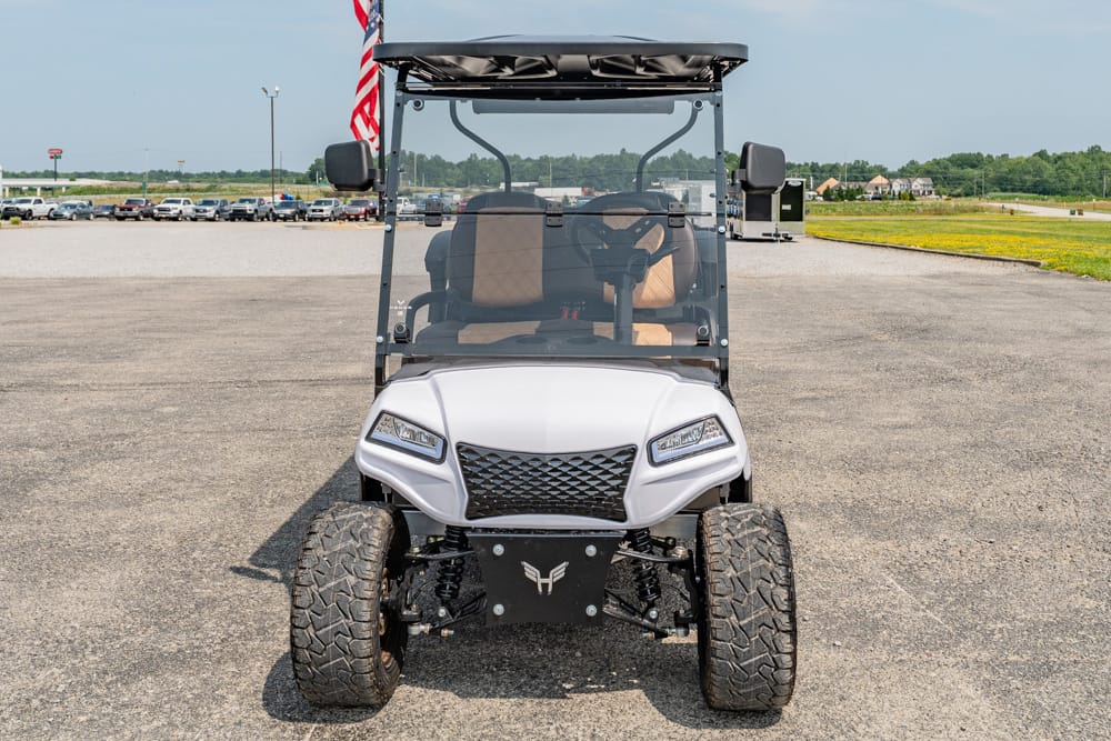Front view of a white off-road golf cart with beige seats and large tires, parked on a wide paved lot with a flag nearby.