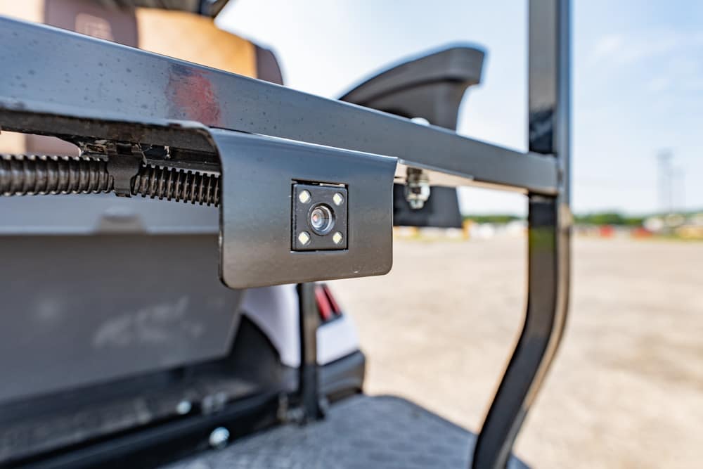 Close-up of a small black square camera mounted on a metal frame of a vehicle, with four LEDs around the lens and an outdoor lot in the background.