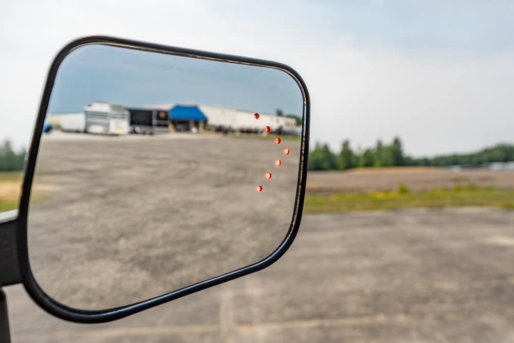 Side mirror reflecting a truck yard with trailers and a blue canopy in the distance.