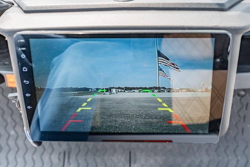 Car's rearview camera display with colored guide lines (green, yellow, red) over a parking lot and an American flag in the distance.