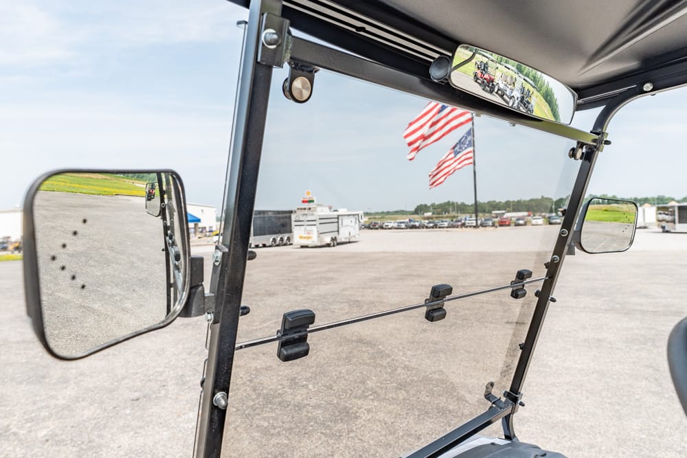Interior view from a vehicle cab looking out at a large parking lot with trailers and American flags in the distance.