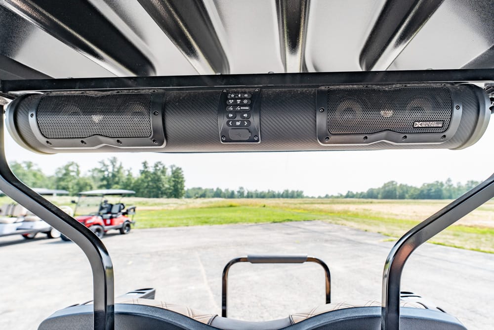 Ceiling-mounted speaker bar with a central control panel inside a golf cart, outdoors in a parking area.