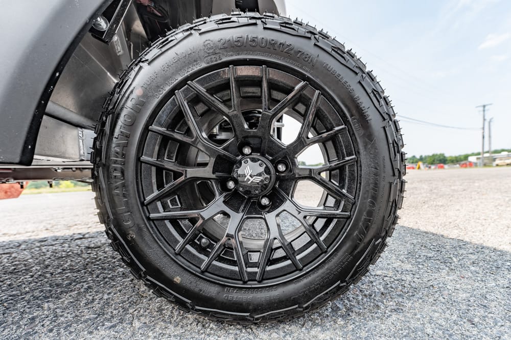 Close-up of a black alloy wheel and rugged tire on a vehicle, parked on a gravel surface.