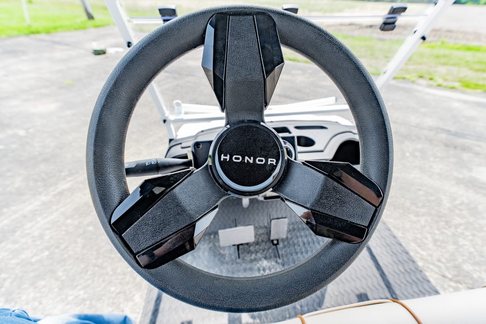 Close-up of a black steering wheel with a circular center emblem reading 'HONOR' on a boat or vehicle dashboard, outdoors on a paved area.