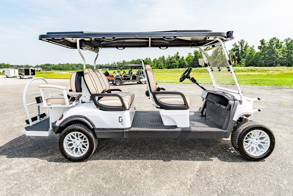 White golf cart with beige seats and a canopy, parked on a paved lot with a grassy field in the background.