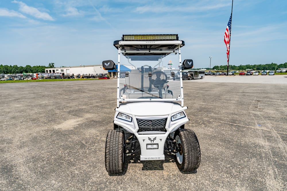 Front view of a white off-road golf cart with rugged dual tires, windshield, and a rooftop light bar in a large paved lot with an American flag nearby.