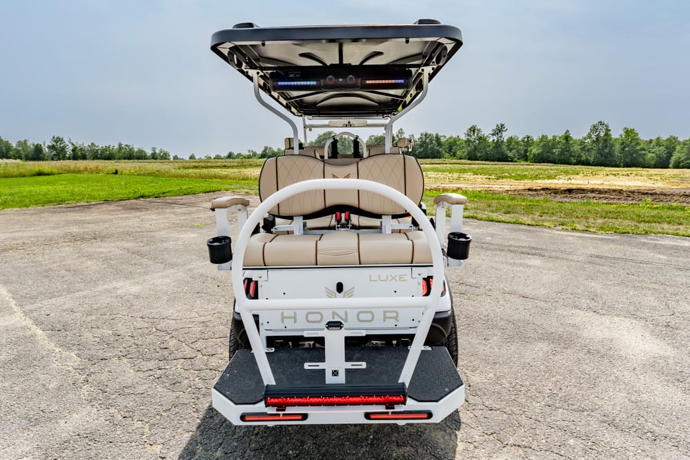 Rear view of a beige luxury golf cart with a canopy, labeled HONOR, parked on a paved surface with fields in the background.