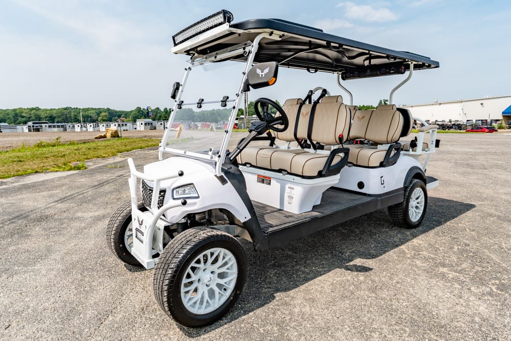 White multi-seat shuttle golf cart with beige seats and roof-mounted lights on a paved tarmac, likely for events or airport use.