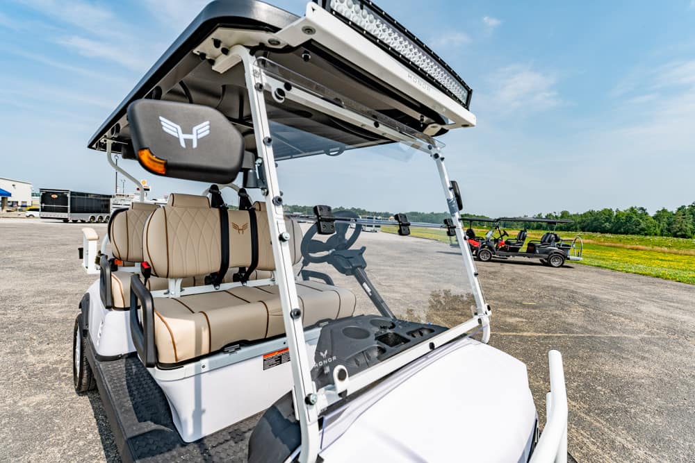 White golf cart with beige leather seats parked on a tarmac, equipped with a roof rack and LED light bar, seen from the front-left side.