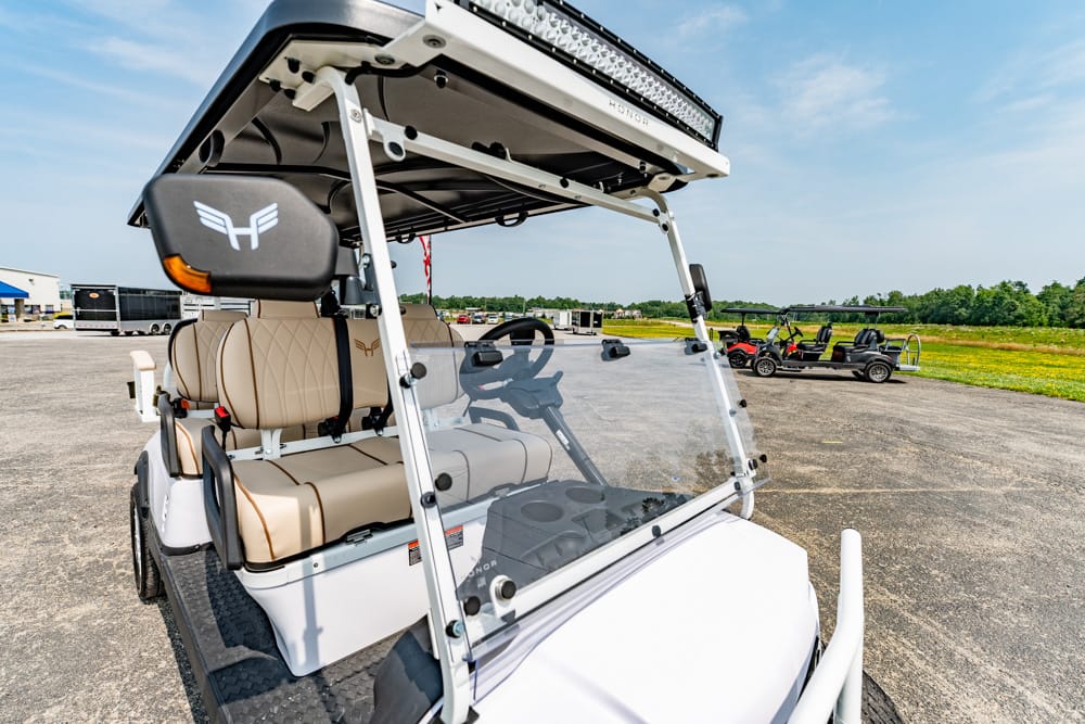 White golf cart with tan seats and a roof-mounted LED light bar, parked on a sunny tarmac with other carts in the distance.