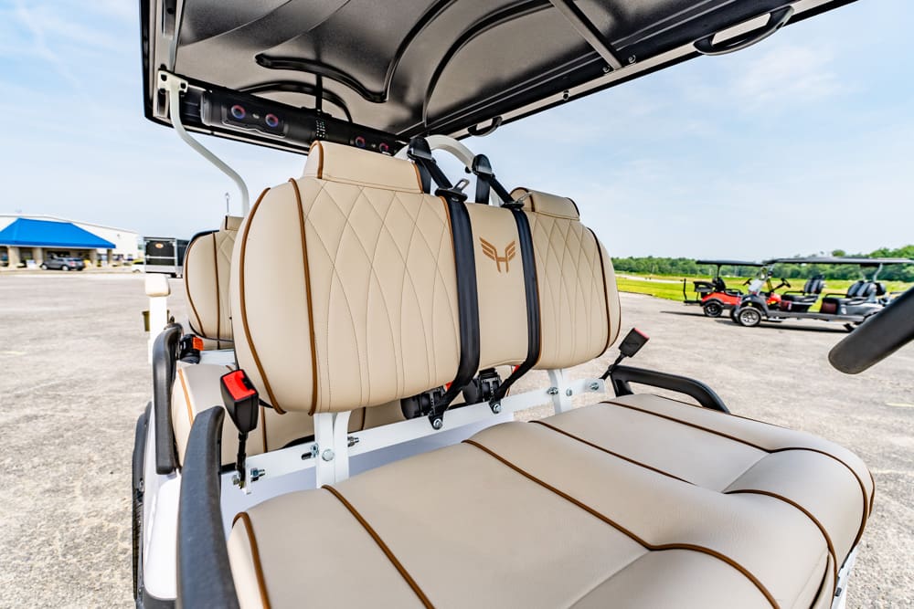 Beige quilted golf cart seats with logo, viewed from the side in an outdoor parking area under an open canopy.