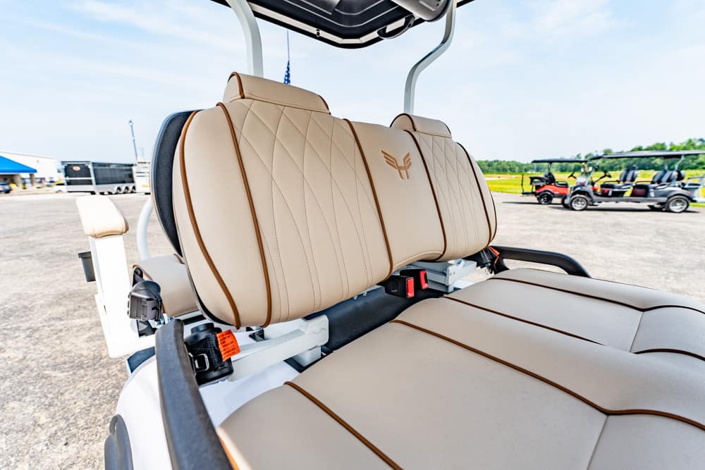 Beige leather captain's seat and bench seat of a golf cart, with brown piping, outdoors on a tarmac lot.