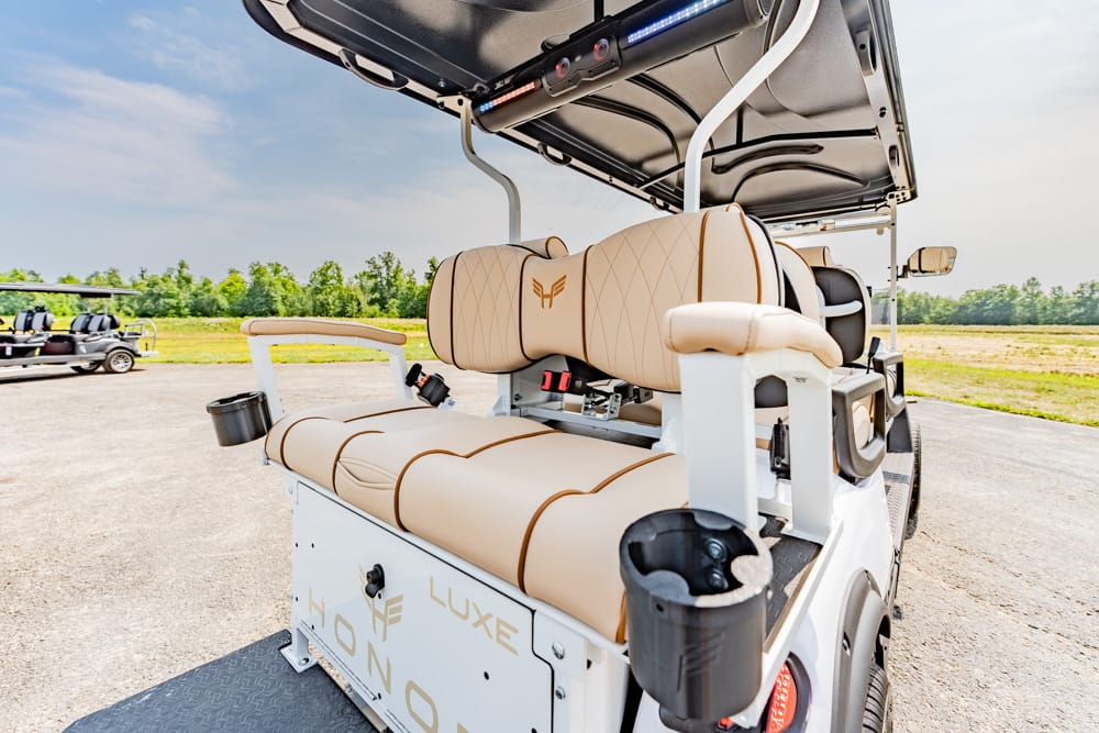 White luxury golf cart with tan leather seats and cup holders, parked on a trailer under an open canopy in a sunny outdoor lot.