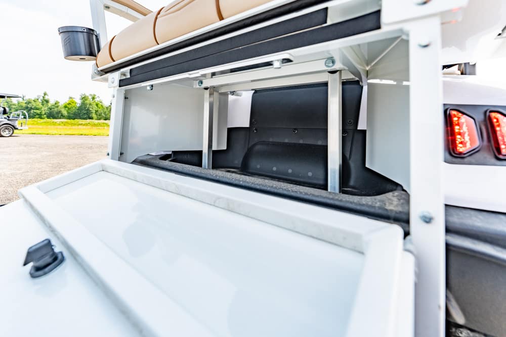 Open rear storage bay of a white utility vehicle, showing metal shelving and a tan cushion inside outdoors on a sunny day.