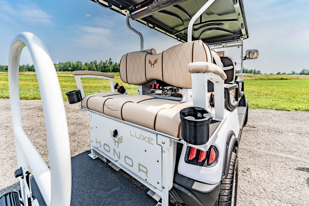 Rear view of a white golf cart with tan leather bench seats and cup holders, on a sunny open field road.
