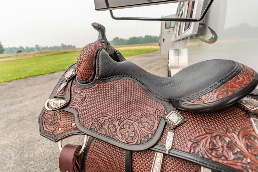 Close-up of a brown, tooled leather Western saddle with floral patterns outdoors, background shows a grassy field and aircraft.