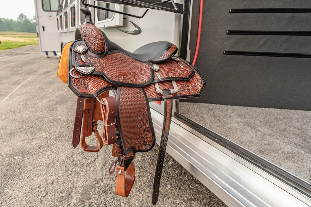 Ornate brown western saddle with decorative tooling and leather straps hanging from a horse trailer doorway on a gravel lot