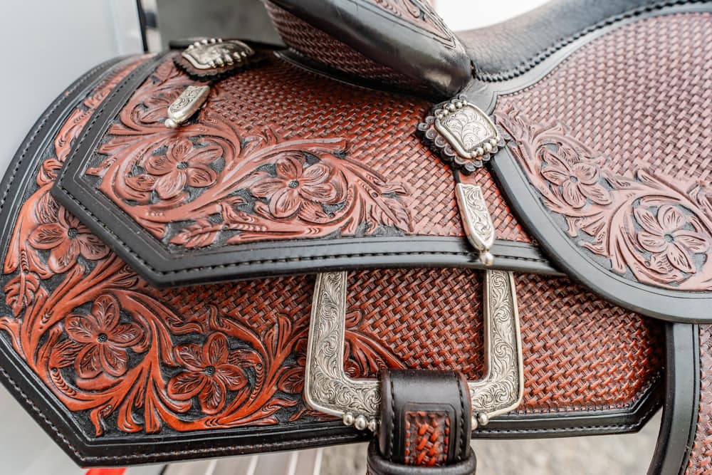 Close-up of red-brown tooled leather horse saddle with floral designs, silver conchos, and a large engraved buckle.