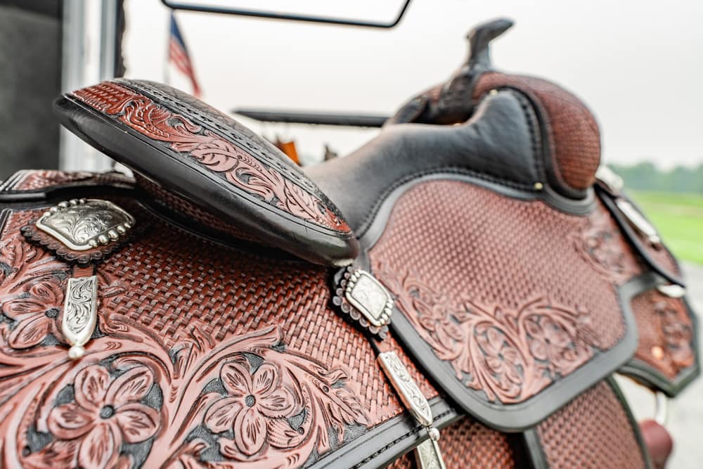 Close-up of a brown tooled leather Western saddle with silver conchos and floral patterns on a horse's back.