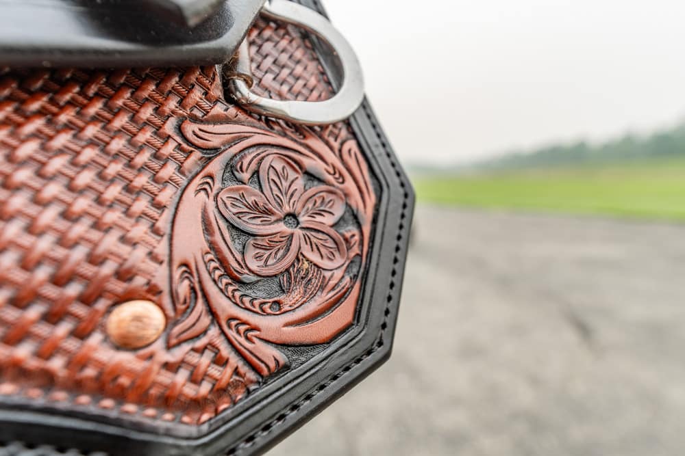 Close-up of brown tooled leather saddle with a floral design and metal ring, outdoors on a blurred landscape.