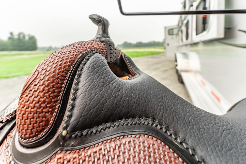 Close-up of a brown and black leather Western saddle, showing detailed stitching, with a horse trailer in the background.