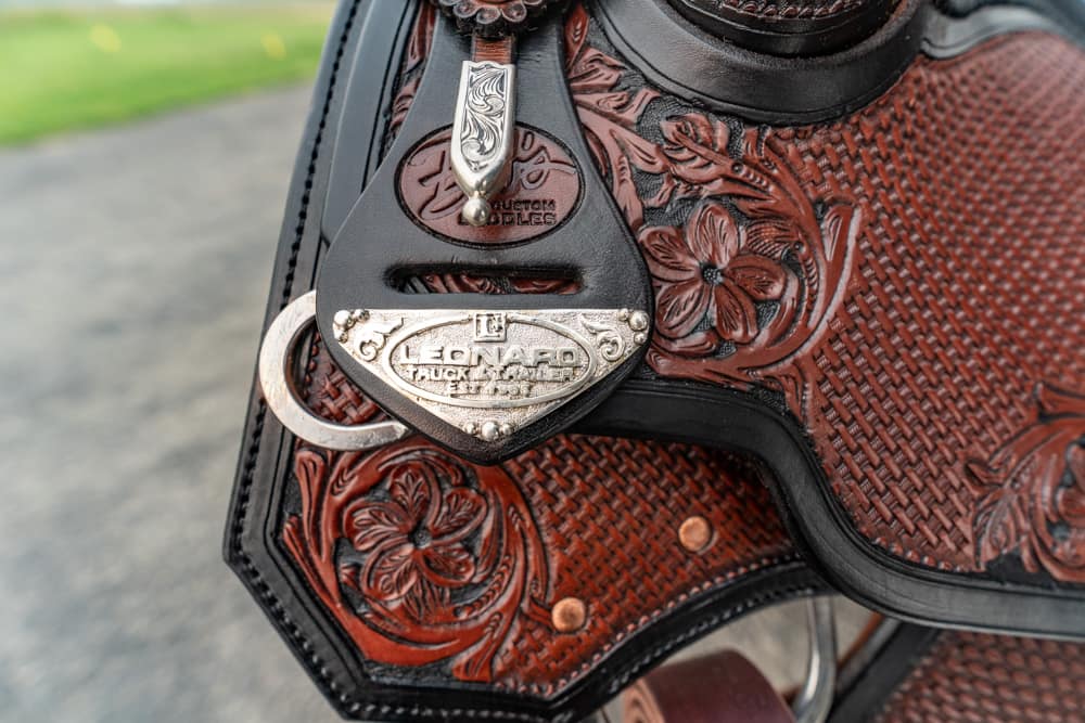 Close-up of decorative leather Western saddle with floral tooling and a silver Leonard buckle on a strap.