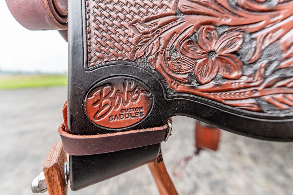 Close-up of a carved leather western saddle with a Boos Custom Saddles badge and decorative floral tooling.