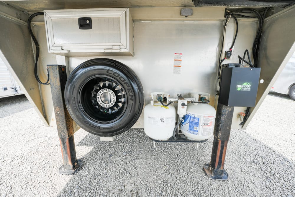 Underbelly of a trailer with a mounted spare tire on the left, two propane tanks and hoses in the center, and a grey utility box labeled Bigfoot on the right, all resting on metal support legs over a gravel surface.