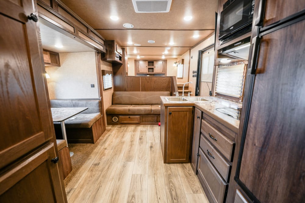 Interior of a wood-toned RV: kitchen with sink and island, dinette booth, and living area at rear.