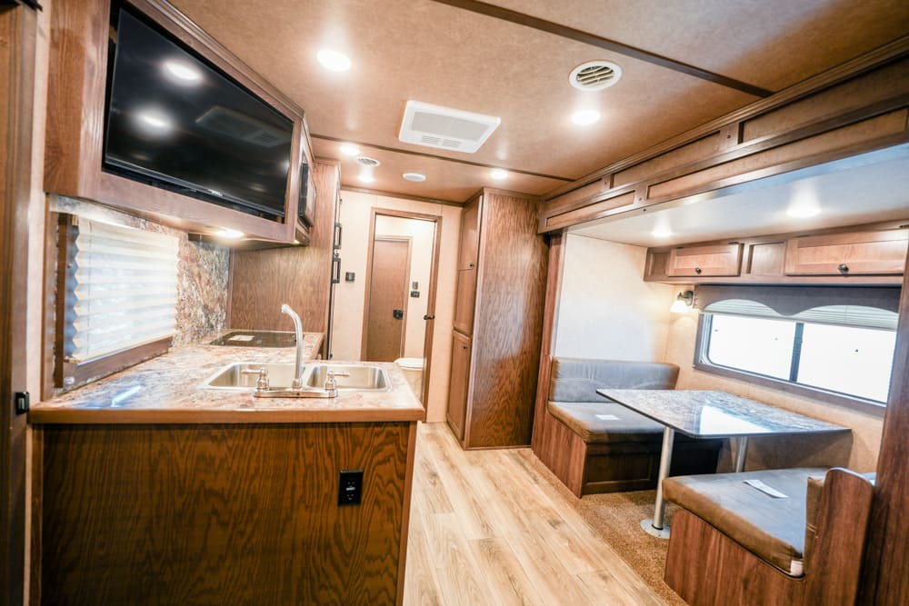 Interior of a compact RV kitchen and dinette: wood cabinets, granite-style counters, double sink, and a seating table along a windowed wall