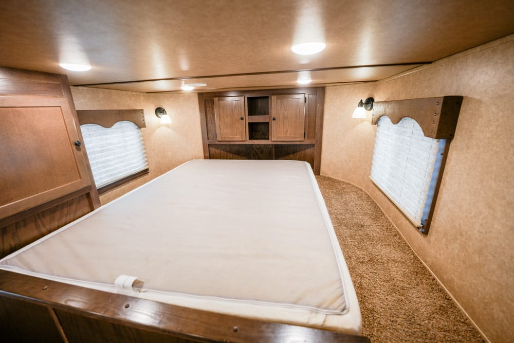 Interior of a camper bedroom with a large central bed, wood cabinetry at the head, and two windows with blinds on either side; warm overhead lighting.