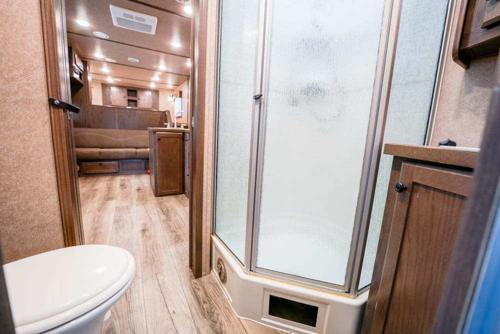 Interior of an RV bathroom with a frosted-glass shower enclosure, toilet in the foreground, and wood cabinetry.