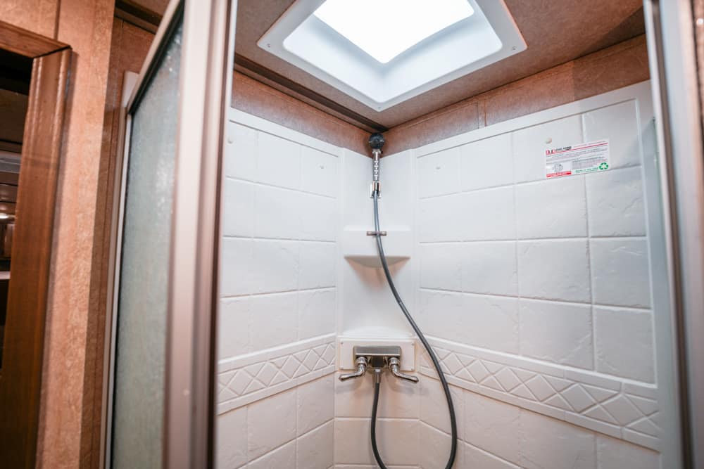 Shower stall with white tiled walls, built-in corner shelf, and a handheld showerhead connected to a dual-handle faucet; skylight above letting light in.