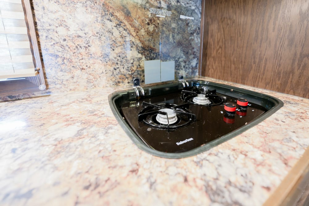 Three-burner gas cooktop set into a speckled granite countertop with red control knobs nearby a wooden wall cabinet into the counter edge