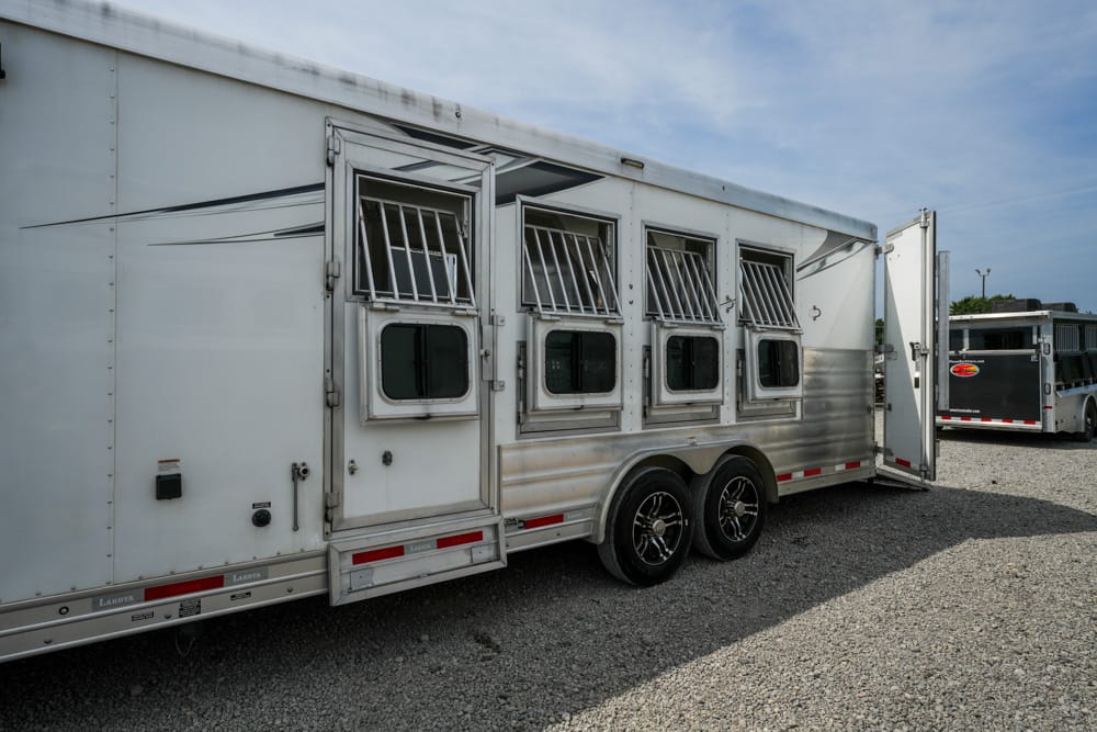 White horse trailer with four ventilated stalls and an open rear ramp in a gravel lot, another trailer visible in the background under a blue sky.