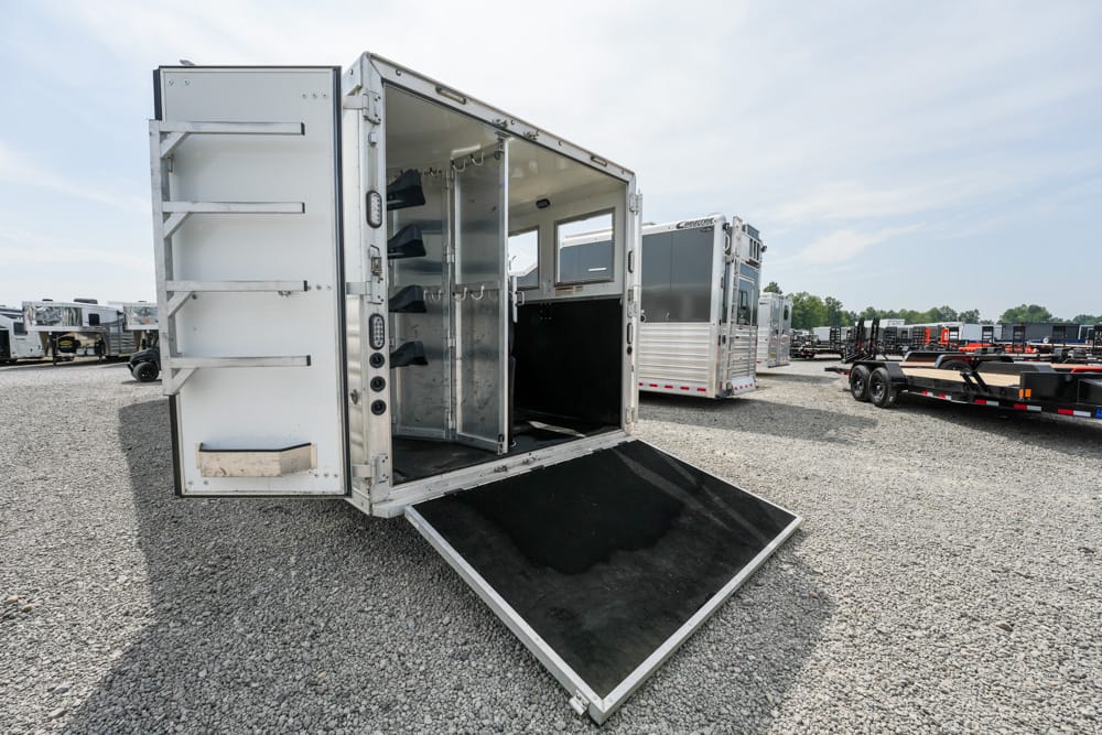Open aluminum utility trailer with ramp deployed on a gravel lot; interior storage shelves and hooks visible on the sides.