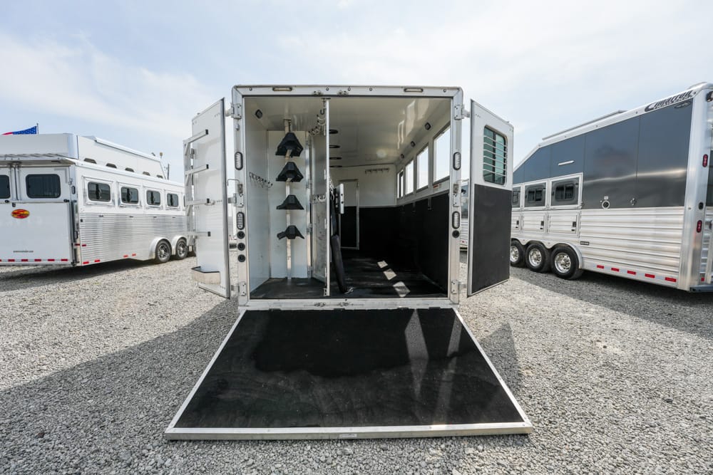 Open horse trailer with a lowered loading ramp, showing stalls, partitions, and tack hooks inside the trailer.