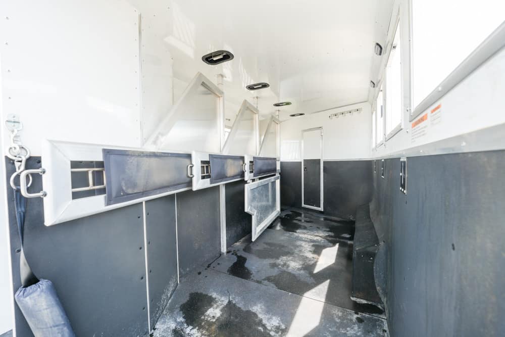 Interior of a horse trailer with dark blue lower walls and white upper sections; several empty stalls with metal dividers along the left side and windows on the right, lit by natural light.